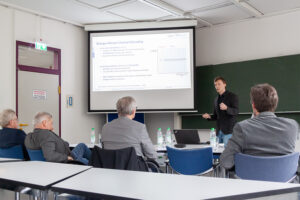 mea presenting in front of beamer and 4 men sitting on tabler