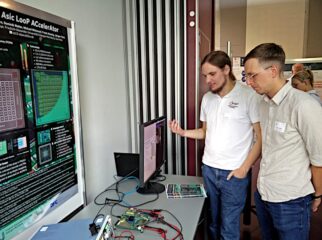 Two person infront of a table with a monitor and a whiteboard