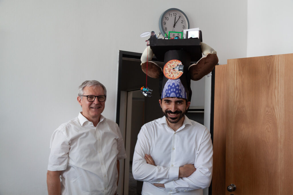 Two smiling men in front of a wall. The one on the right wears a doctoral hat